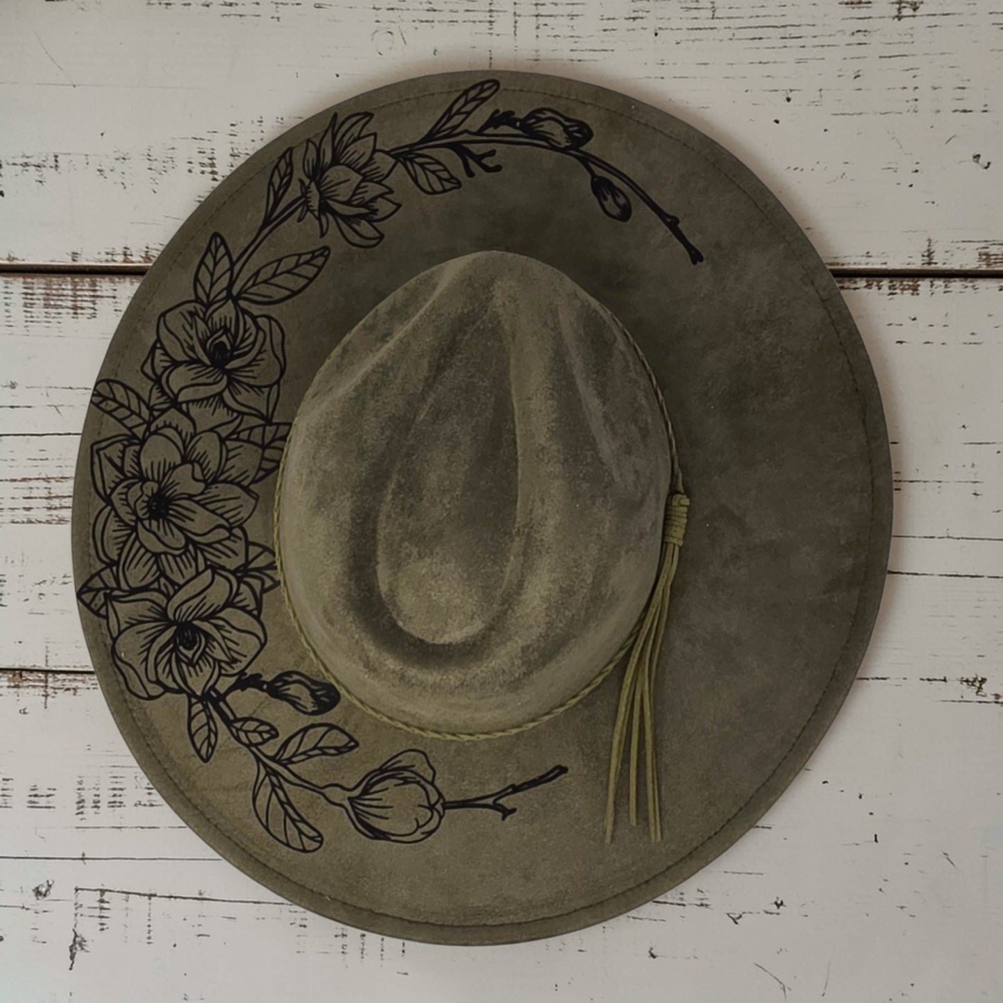 Elkhorn Designs sage green vegan suede wide-brim hat with intricate laser-engraved gardenia floral design and tassel.