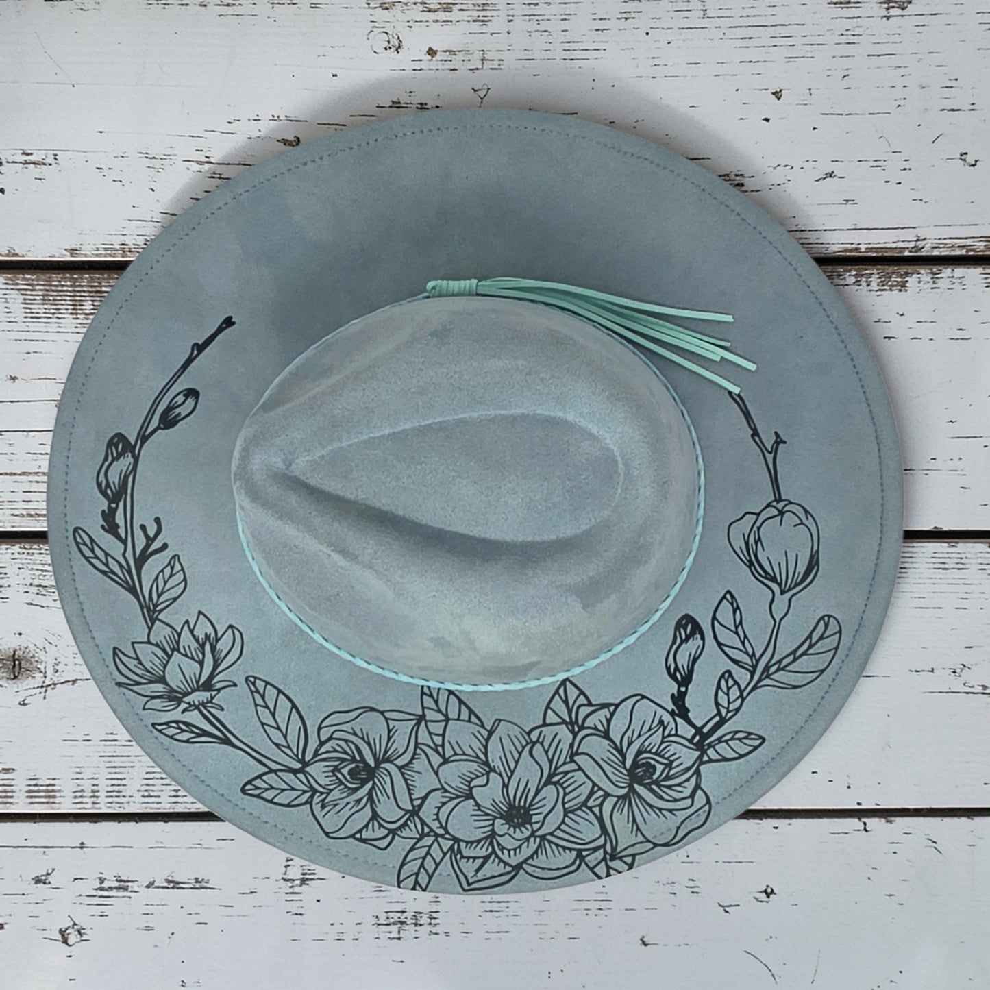 Elkhorn Designs sea glass green vegan suede wide-brim hat with laser-engraved gardenia floral pattern and tassel detail.