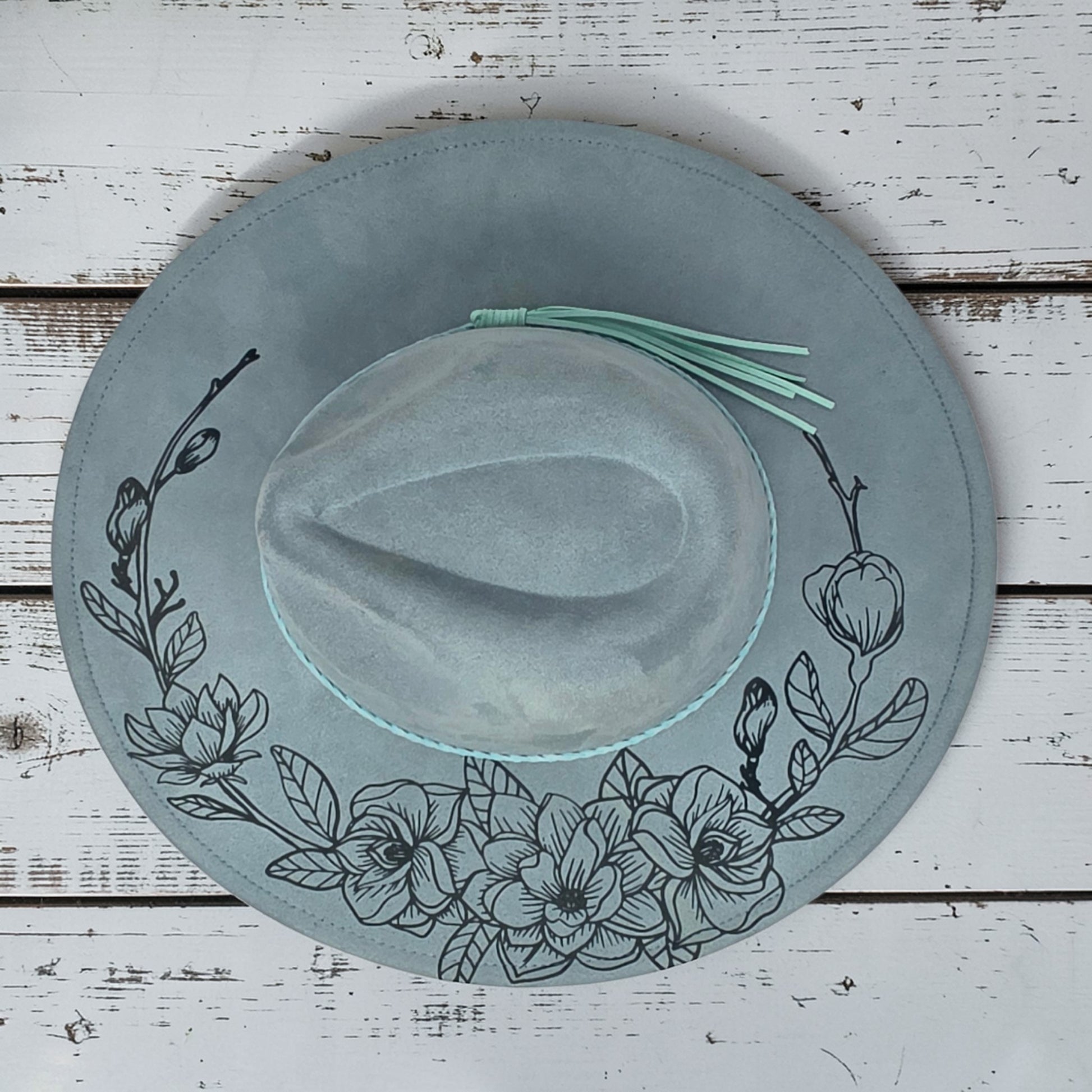 Elkhorn Designs sea glass green vegan suede wide-brim hat with laser-engraved gardenia floral pattern and tassel detail.